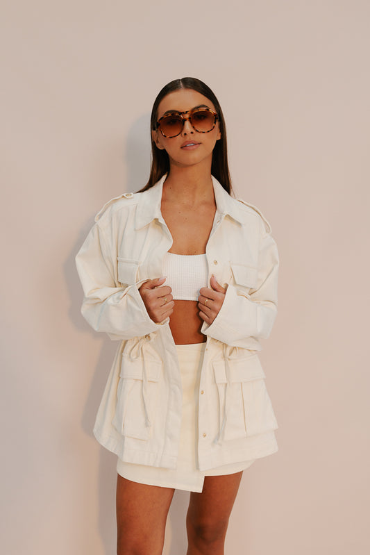 A female model wearing a cream utility jacket with a relaxed fit, oversized silhouette, and elasticated waistband. She is also wearing a white top and beige skirt, accessorized with sunglasses.
