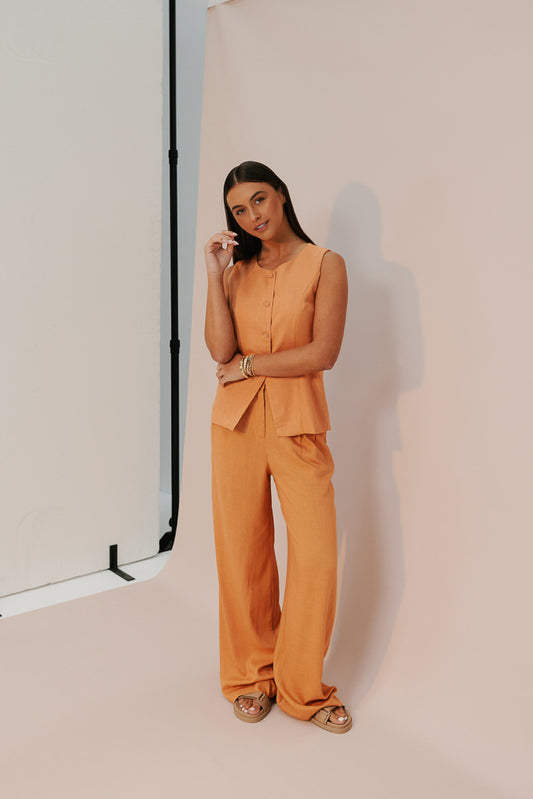 A model wearing a rust orange linen blend tailored pant with a relaxed fit, featuring a mid waist fit with darts, side slip pockets and a stretch elastic to the back.