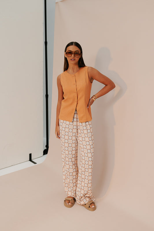 A model wearing a rust orange sleeveless vest with a round neckline and fabric-covered buttons, paired with white pants adorned with a geometric pattern.